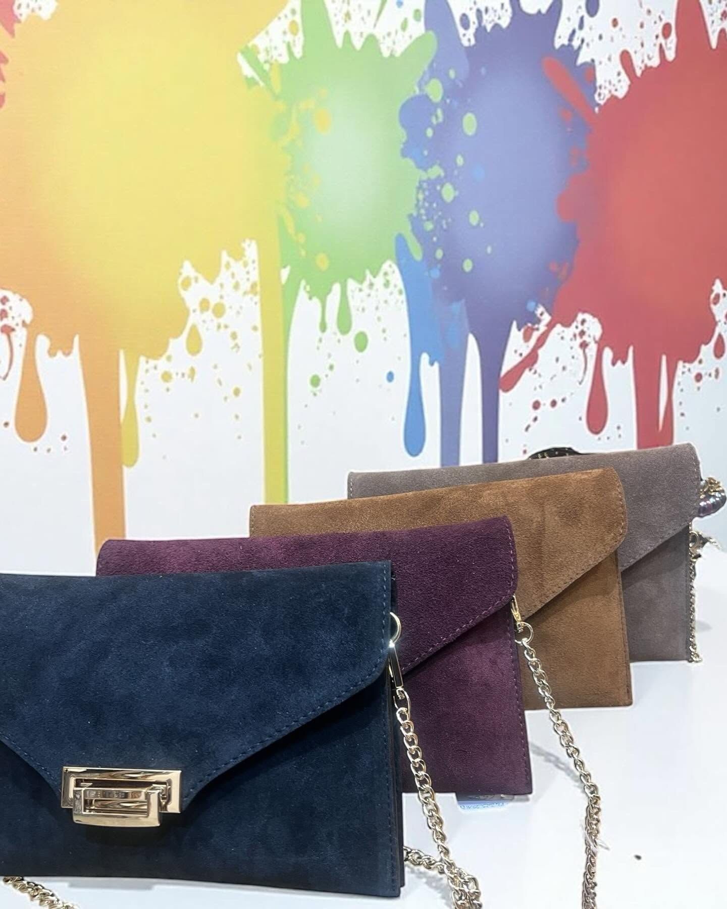 Quattro pochette in camoscio blu, prugna, cuoio e grigio con catenella dorata davanti a un murale di schizzi di vernice
