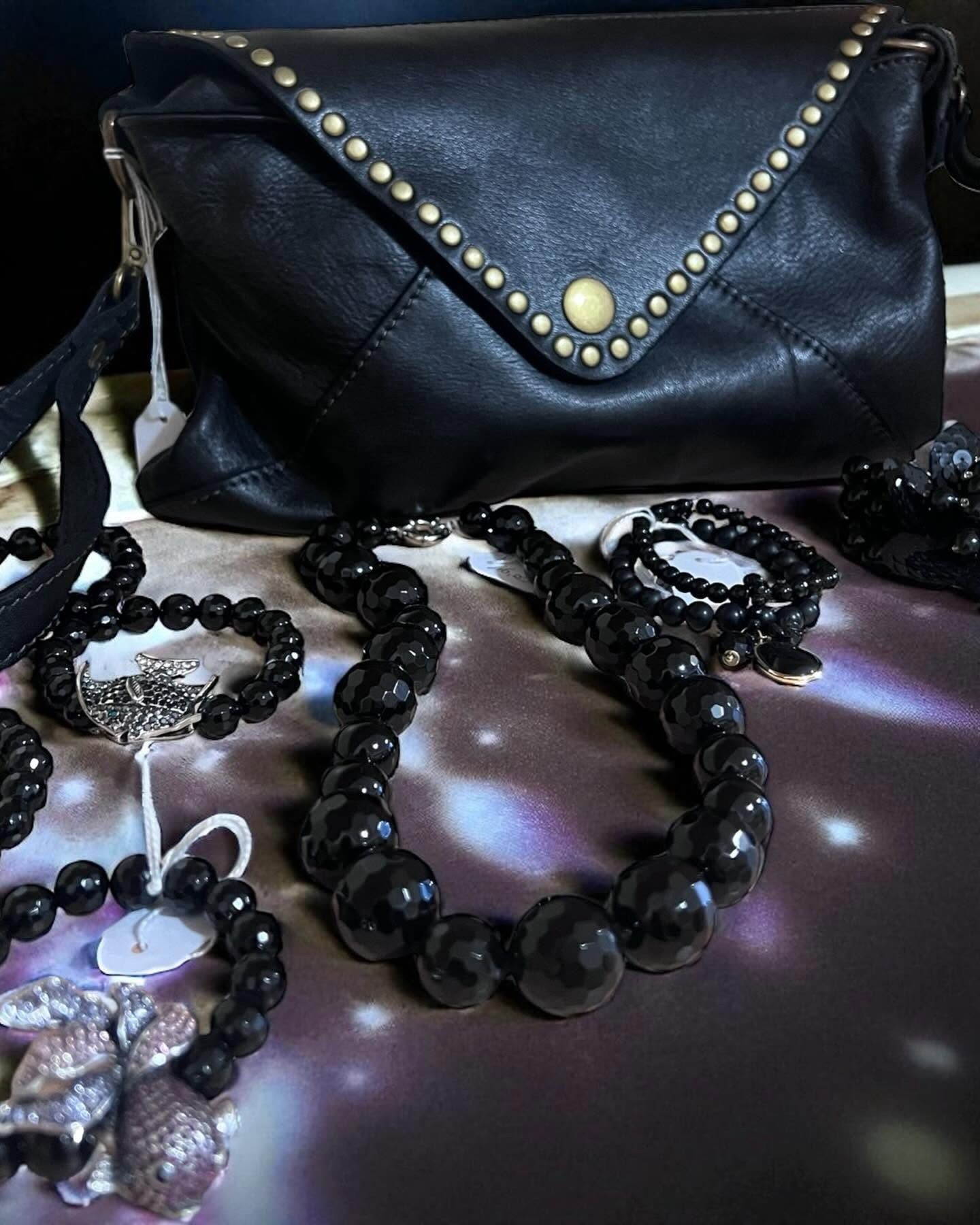 Borsa in pelle nera con borchie dorate, collane e bracciali di perle nere sfaccettate e spille argentate su tessuto viola